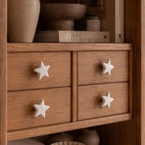 Puede incluir: Armario de madera clara con cuatro cajones, cada uno adornado con un pomo blanco en forma de estrella. El armario está decorado con objetos, incluyendo un jarrón, un cuenco y un libro con el texto "AD at 100 LIVE BEAUTIFULLY".
