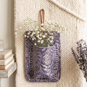 Może przedstawiać: Fioletowa ceramiczna kieszeń ścienna z kwiatowym wzorem, wypełniona białymi kwiatami. Kieszeń ma skórzaną pętlę do zawieszenia. Tło to kremowa, teksturowana tkanina.