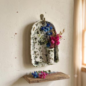 May include: A wall-mounted ceramic vessel with a white base and black speckled pattern, featuring an arched shape and a small integrated bud vase. The vessel holds a bouquet of dried blue, pink, and gold flowers, hanging from a metal hook.