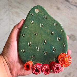 May include: A green ceramic wall hanging shaped like a prickly pear cactus pad with a hole at the top for mounting. The surface features small V-shaped indentations and five textured, bright orange-red flower blossoms along the bottom edge.