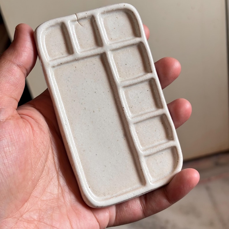 Pu&ograve; includere: Una tavolozza rettangolare in ceramica bianco sporco con pi&ugrave; pozzetti per mescolare i colori. La tavolozza ha una consistenza leggermente macchiata e una crepa vicino alla parte superiore. La tavolozza &egrave; tenuta in una mano.