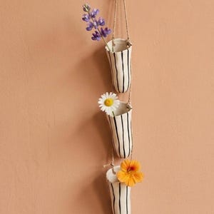 May include: A vertical wall-hanging set of three off-white ceramic bud vases, each featuring hand-painted black vertical stripes. The vases are connected by thin twine and suspended from a single hook, holding purple, white, and orange flowers.