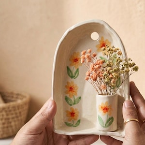 Può includere: Un supporto da parete in ceramica color bianco sporco a forma di arco, decorato con fiori dipinti a mano in giallo e arancione con foglie verdi. Un piccolo vaso cilindrico è fissato sul davanti, contenente un bouquet di fiori secchi rosa e verdi.