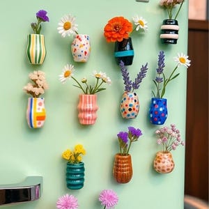 May include: A collection of miniature decorative vases in various colors and patterns, each holding a small bouquet of flowers. The word "VASE" is spelled out in silver letters above the vases, which are attached to a light green surface.