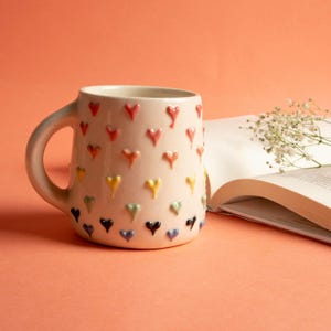 May include: Cream-colored ceramic mug adorned with raised, colorful heart-shaped designs. The hearts are in a rainbow of colors. The mug has a rounded handle and sits next to an open book and sprigs of white flowers.