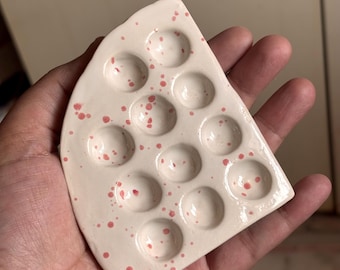 Pode incluir: Uma paleta de pintura de cerâmica em forma de fatia de queijo. A paleta esbranquiçada tem salpicos cor-de-rosa e 12 poços circulares para tinta. A paleta é segurada em uma mão.
