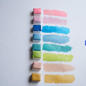 May include: A collection of watercolor paints in various colors, including pink, blue, green, and yellow. Each color is displayed as a small square of paint next to a corresponding brushstroke on white paper. The image showcases the vibrant hues and textures of the watercolor paints.