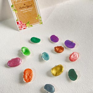 May include: A selection of small, oval watercolour paint samples in shades of green, purple, orange, and blue. The samples are scattered on a white surface, with a card that reads "My Tiny Journalie" and "Handmade Watercolour Journals & Paints."