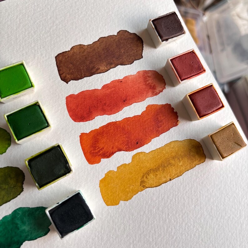 May include: Watercolor paint set with various colors, including green, brown, red, and yellow. The paint is in small square pans, with swatches of each color painted on white paper. The colors range from dark to light shades.