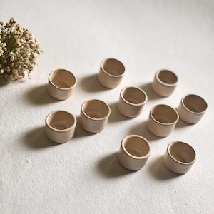 May include: Ten small, round, beige ceramic bowls arranged on a white surface. The bowls are all the same size and shape, and they have a slightly textured finish.