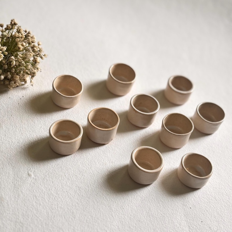 May include: Ten small, round, beige ceramic bowls arranged on a white surface. The bowls are all the same size and shape, and they have a slightly textured finish.