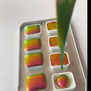 K&ouml;nnte beinhalten: Nahaufnahme einer Metallschale mit Aquarellfarben. Die Pfannen sind mit leuchtenden, irisierenden Farben in Gr&uuml;n-, Gelb- und Rott&ouml;nen gef&uuml;llt. Ein Strahl gr&uuml;ner Farbe wird in eine der Pfannen gegossen.