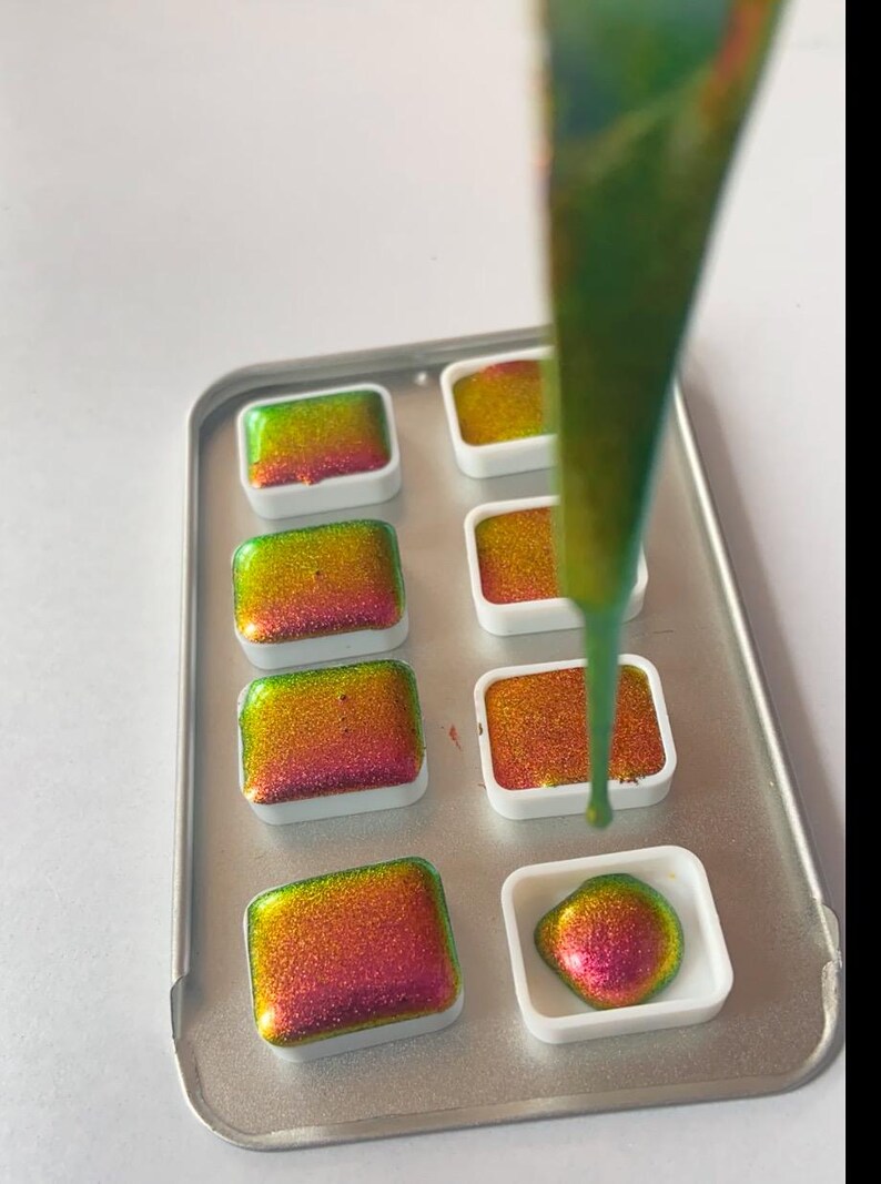 K&ouml;nnte beinhalten: Nahaufnahme einer Metallschale mit Aquarellfarben. Die Pfannen sind mit leuchtenden, irisierenden Farben in Gr&uuml;n-, Gelb- und Rott&ouml;nen gef&uuml;llt. Ein Strahl gr&uuml;ner Farbe wird in eine der Pfannen gegossen.