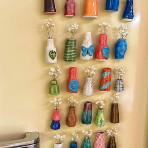 Può includere: Una collezione di piccoli vasi in ceramica colorati, ognuno con un rametto di fiori bianchi secchi. I vasi presentano varie forme e disegni, tra cui strisce, fiocchi e motivi floreali. Sono attaccati a una superficie color crema.
