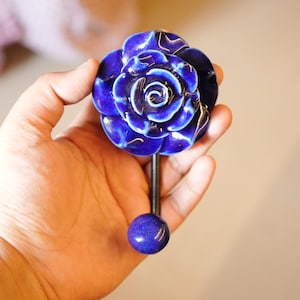Puede incluir: Un gancho con forma de rosa de cerámica azul, con un tallo oscuro y una base redonda azul. La rosa tiene pétalos detallados y un acabado brillante. El gancho se sostiene en una mano sobre un fondo neutro.