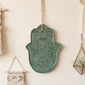 May include: A sage green ceramic hamsa hand wall hanging featuring intricate embossed geometric patterns, a central mandala design, and a small eye motif. The piece is suspended by a thin brown twine cord through a hole at the top centre.