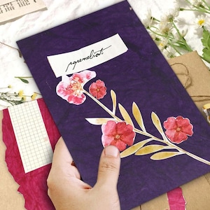 May include: A purple notebook with pressed flowers and the word "agremeliorati" on a white label. The notebook features a floral design with red flowers and yellow leaves. The notebook is held by a hand.