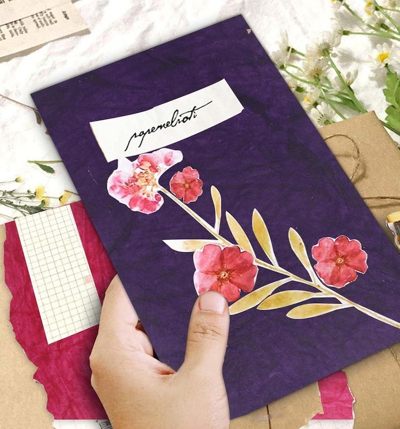May include: A purple notebook with pressed flowers and the word "agremeliorati" on a white label. The notebook features a floral design with red flowers and yellow leaves. The notebook is held by a hand.