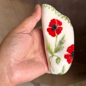 Może przedstawiać: Zakrzywiony wazon ceramiczny w kolorze złamanej bieli, ozdobiony ręcznie malowanymi czerwonymi makami z czarnymi środkami oraz zielonymi łodygami i liśćmi. Krawędź wazonu zdobi delikatny wz&oacute;r w zielone kropki wzdłuż brzegu białej powierzchni.