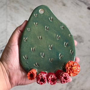 May include: A teardrop-shaped green ceramic wall hanging with a glossy finish and small V-shaped indentations across the surface. The bottom edge features five textured, bright orange and red flower-like protrusions. A circular hole is at the top.