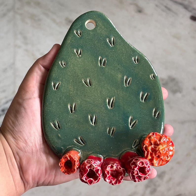 May include: A teardrop-shaped green ceramic wall hanging with a glossy finish and small V-shaped indentations across the surface. The bottom edge features five textured, bright orange and red flower-like protrusions. A circular hole is at the top.