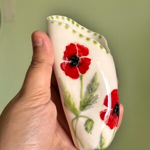 Może przedstawiać: Biały ceramiczny wazon ścienny z błyszczącym wykończeniem, ozdobiony ręcznie malowanymi czerwonymi makami z czarnymi środkami i zielonymi łodygami. G&oacute;rna krawędź jest udekorowana serią małych zielonych kropek, a przedmiot ma wydłużony, zakrzywiony kształt.