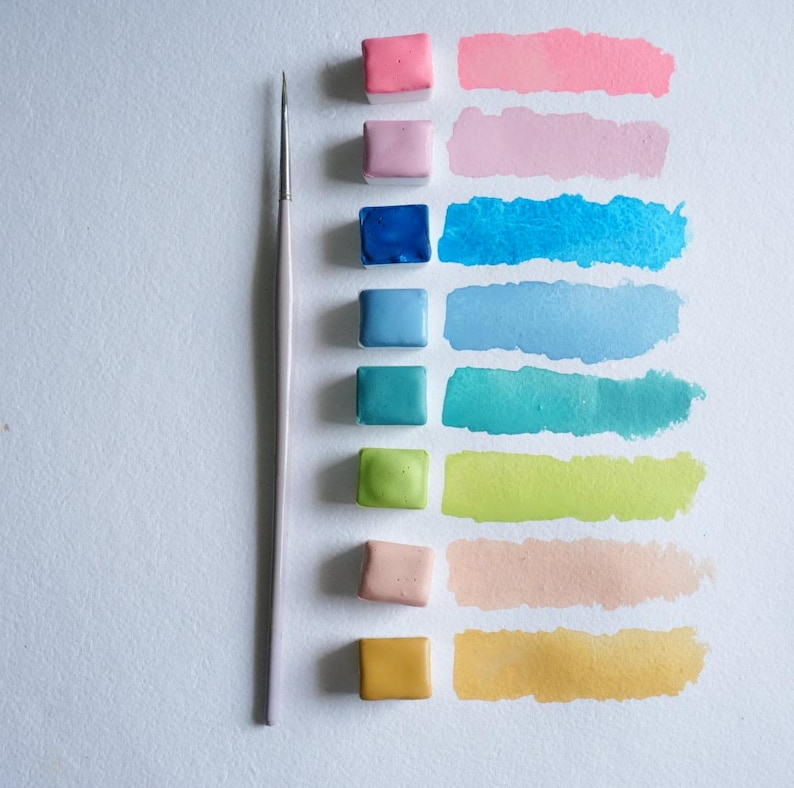 May include: A watercolor paint set with a variety of colors, including pink, blue, green, and yellow. Each color is displayed as a square of solid paint and a corresponding brushstroke. A thin paintbrush is positioned vertically.