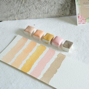 May include: A collection of six watercolor paint pans in various shades of peach, yellow, pink, and brown, arranged above corresponding color swatches on white paper. The paints are in small, square, white containers. The swatches are painted in diagonal stripes.