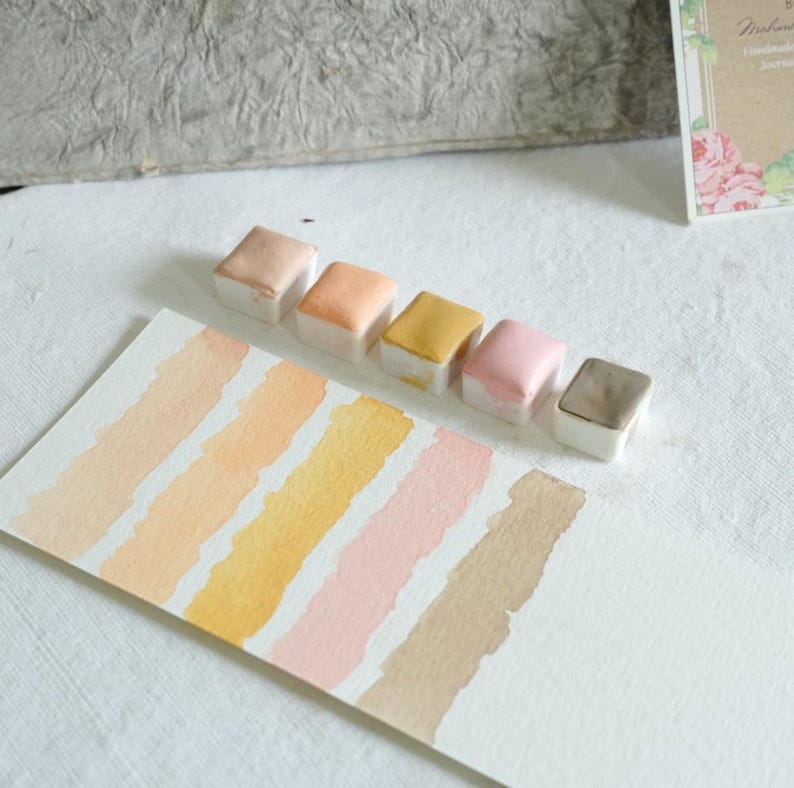 May include: A collection of six watercolor paint pans in various shades of peach, yellow, pink, and brown, arranged above corresponding color swatches on white paper. The paints are in small, square, white containers. The swatches are painted in diagonal stripes.