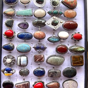 Puede incluir: Exhibición de anillos con piedras preciosas con engastes plateados. Los anillos presentan una variedad de formas y colores de piedras, incluyendo azul, verde, rojo y marrón. Los anillos están dispuestos en un patrón de cuadrícula.