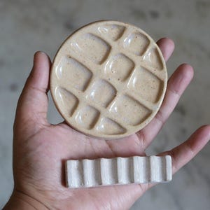 May include: A round, speckled beige ceramic paint palette with a grid of recessed wells and a separate rectangular, off-white ceramic brush rest featuring a series of parallel grooves for holding multiple painting tools securely.