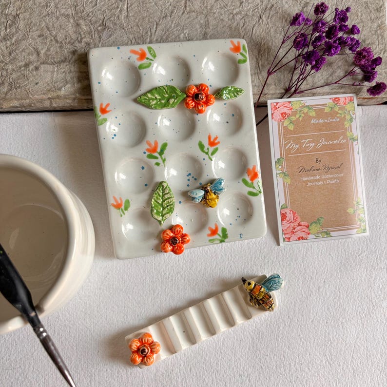 May include: A white ceramic paint palette with 16 wells, decorated with orange flowers, green leaves, and bees. The palette is handmade and comes with a small white ceramic palette knife.