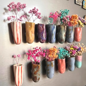 May include: A collection of small, ceramic vases in various colours, each holding dried flowers. The vases are mounted on a wall, showcasing a range of hues from pink and orange to blue and purple. The vases are approximately 10 cm tall.
