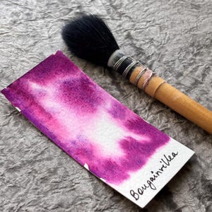 May include: A watercolour brush with a wooden handle and black bristles lies next to a watercolour swatch. The swatch is a rectangular paper with a blend of pink and purple hues, with the word "Bougainvillea" written on it.