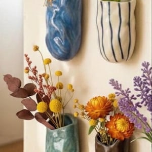 May include: A collection of wall-mounted ceramic vases in various colors, each holding different dried flowers. The vases are in shades of blue, green, white, and brown. The flowers include daisies, yellow pom-poms, and orange blooms.