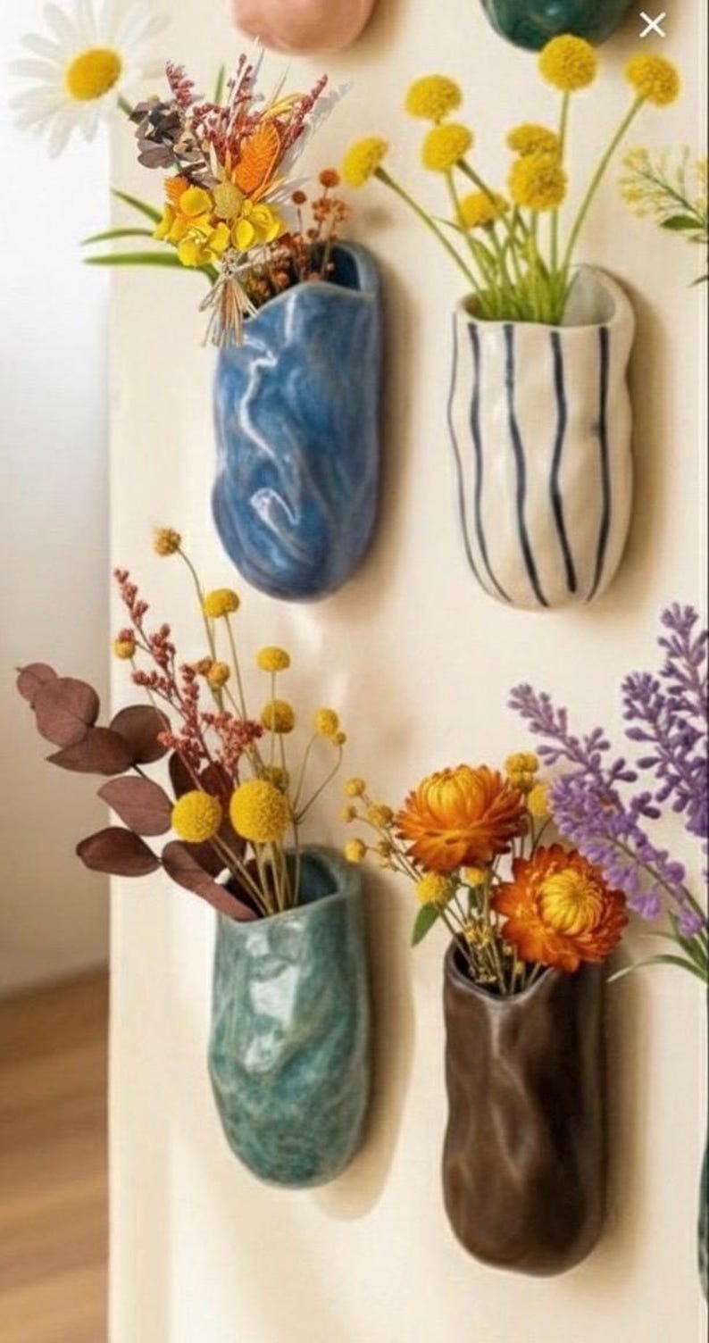 May include: A collection of wall-mounted ceramic vases in various colors, each holding different dried flowers. The vases are in shades of blue, green, white, and brown. The flowers include daisies, yellow pom-poms, and orange blooms.
