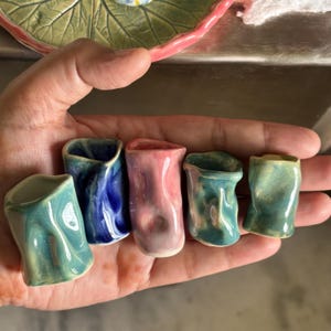 May include: A collection of six small, handmade ceramic shot glasses in various colors, including teal, blue, and pink. Each glass has a unique, organic shape and a glossy finish. The shot glasses are held in a hand, showcasing their size and detail.