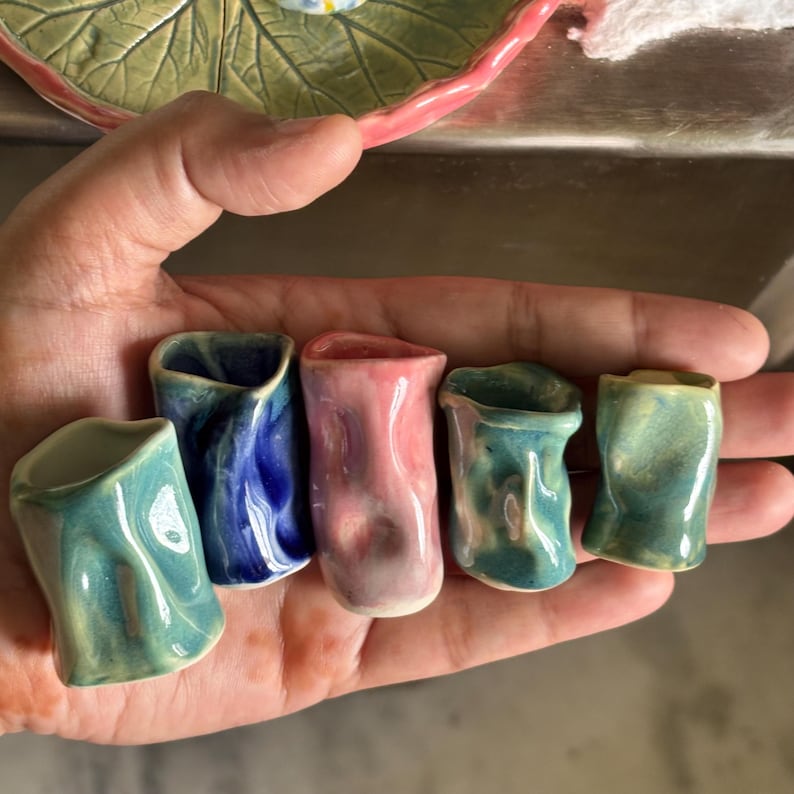 May include: A collection of six small, handmade ceramic shot glasses in various colors, including teal, blue, and pink. Each glass has a unique, organic shape and a glossy finish. The shot glasses are held in a hand, showcasing their size and detail.