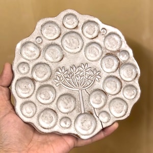 May include: A handmade, off-white ceramic palette with multiple circular wells of varying sizes. A stylized botanical design is centered on the palette. The palette has a rustic, organic shape and a crack along the edge.