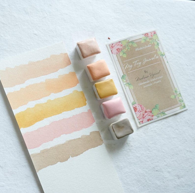 May include: A watercolor palette with six square paint blocks in shades of beige, peach, yellow, pink, and gray. A watercolor swatch card displays matching color swatches. A business card with floral accents reads "Tiny Toy Journals" and "Handmade Watercolor Journals & Palettes."