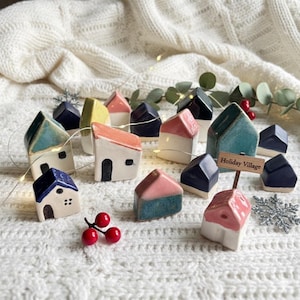 May include: A collection of miniature ceramic houses in various colors, including blue, pink, and yellow, arranged on a white knitted surface. Some houses have a two-tone design. A small wooden sign reads "Holiday Village".