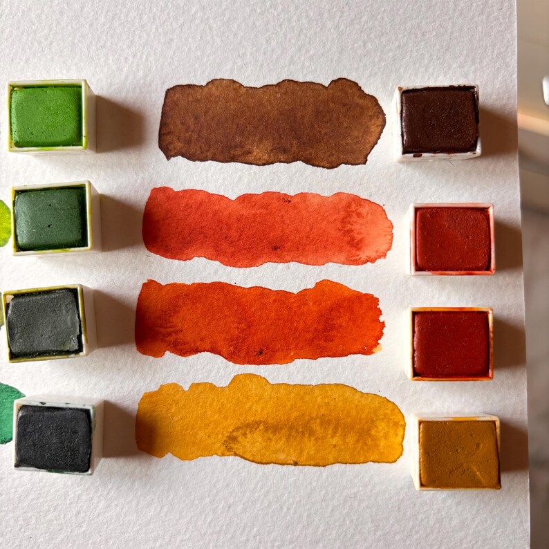 May include: A collection of watercolor paints in various colors, including green, brown, orange, and yellow. Each color is displayed as a solid square and a painted swatch, showcasing the pigment's hue and texture on white paper.