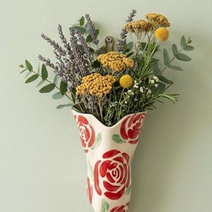 Pode incluir: Um vaso de cerâmica montado na parede com uma base de cor creme e motivos de rosas vermelhas pintados à mão com folhas verdes. O vaso está preenchido com um arranjo de flores secas com lavanda roxa, milefólio amarelo e eucalipto.