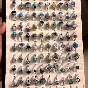 May include: A collection of silver-toned rings with various gemstone settings, including turquoise, amethyst, and tiger's eye. The rings are displayed on a white surface, showcasing a variety of shapes and sizes, suitable for jewelry enthusiasts.