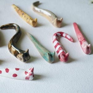 May include: A collection of whimsical ceramic cat-shaped incense holders in various colors and patterns. The holders feature a curved design with a cat head at the end. Colors include yellow, brown, green, red, and white, with some pieces displaying polka dots or stripes.