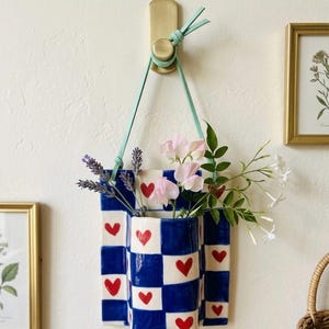 Pode incluir: Um vaso de parede de cerâmica xadrez azul e branco com detalhes de corações vermelhos, pendurado em um gancho de latão com uma alça de couro azul claro. O vaso contém lavanda, ervilhas-de-cheiro rosa e flores de jasmim brancas.
