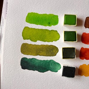 May include: A close-up of a watercolor paint swatch on white paper. The swatches are in shades of green, with four rectangular paint pans of green paint. The paint is arranged in a row, with the swatches below the paint pans.