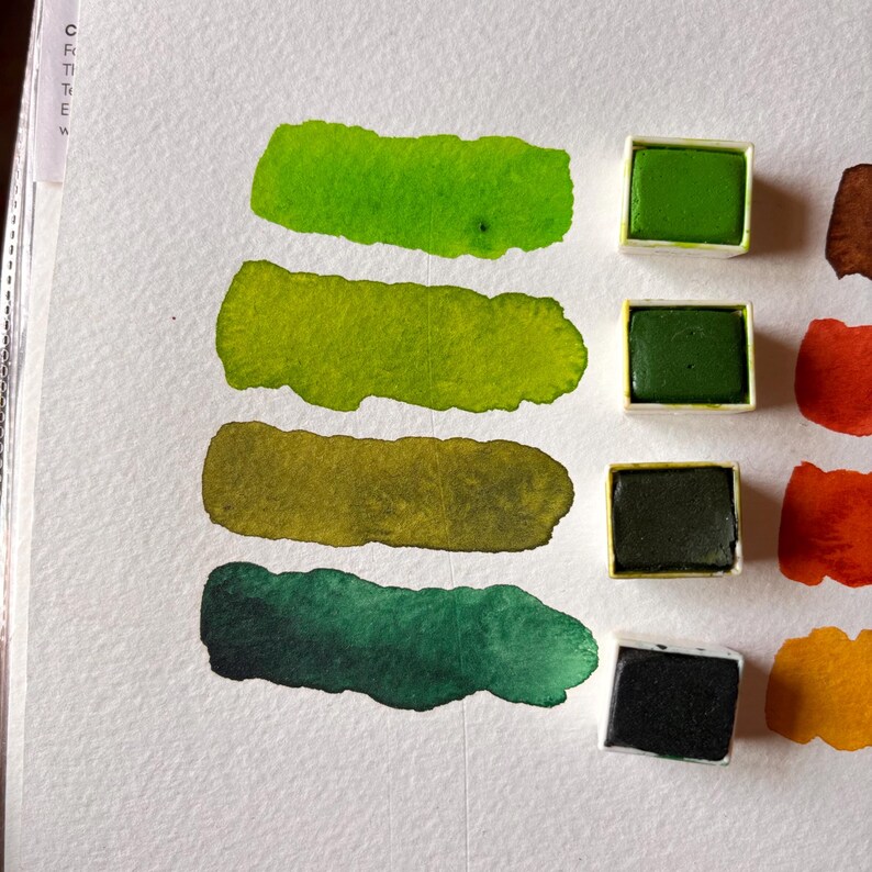 May include: A close-up of a watercolor paint swatch on white paper. The swatches are in shades of green, with four rectangular paint pans of green paint. The paint is arranged in a row, with the swatches below the paint pans.