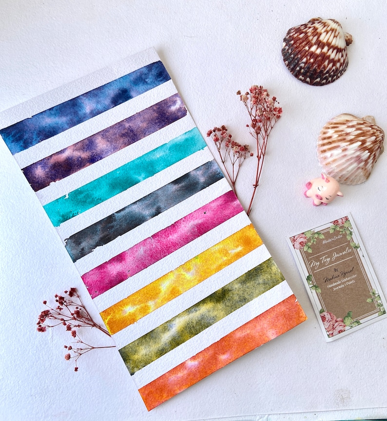 May include: A watercolor painting with horizontal stripes of various colors, including blue, purple, teal, black, pink, yellow, green, and orange. The painting is on white paper, and the stripes are separated by white lines. The image also includes seashells and a small pig figurine.