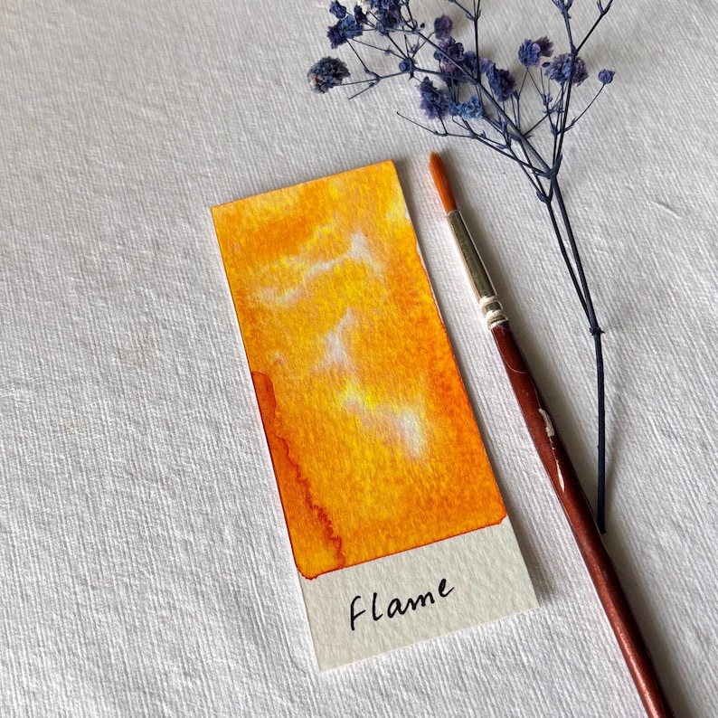 K&ouml;nnte beinhalten: Ein rechteckiger Aquarellfarbmuster in Gelb- und Oranget&ouml;nen, beschriftet mit "Flame". Ein Pinsel mit braunem Griff und ein Zweig getrockneter blauer Blumen liegen neben dem Muster. Das Muster hat einen wei&szlig;en Rand.